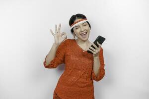 une de bonne humeur asiatique femme portant rouge kebaya et Bandeau, en portant sa téléphone tandis que faire des gestes D'accord signe avec sa des doigts, isolé par blanc Contexte. l'indonésie indépendance journée concept photo