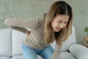 Asie magnifique femme en portant sa inférieur retour tandis que et souffrir de insupportable douleur santé et problèmes, chronique retour douleur, mal au dos dans Bureau syndrome, scoliose, hernie disque, muscle inflammation photo