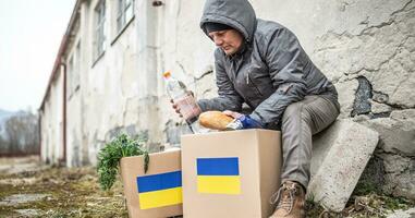 vysne nemecke, slovaquie. Mars 30. 2022. plus âgée homme dans chaud vêtements est assis sur le bord de dérélictionné bâtiment prise pain et l'eau en dehors de humanitaire aide paquet pour Ukraine photo