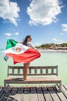 mexicain patriotique femme détient une nationale drapeau permanent sur une en bois banc suivant à le mer photo