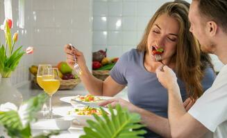 content couple en mangeant salade dans cuisine à maison. Jeune homme donnant fraise à le sien petite amie. photo