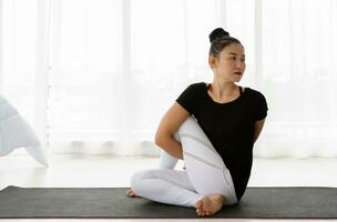 asiatique femmes Faire yoga assise torsion exercice à maison, séance dans Seigneur de le moitié des poissons pose ou ardha matsyendrasana, dans blanc chambre à coucher. travail en dehors portant noir chemise et blanc pantalon. photo