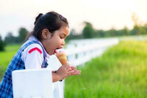 mignonne asiatique fille dans école uniforme en mangeant la glace crème tandis que sur vacances avec famille, et brouillé de vert champ riz la nature arrière-plan, content famille concept photo