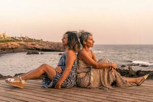 deux charmant milieu vieilli copines séance sur une en bois pont par le mer à le coucher du soleil photo