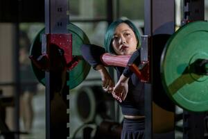 asiatique femme est orienté vers surentraînement syndrome sur entraine toi poids levage en utilisant haltère pour bras et coeur muscle à l'intérieur Gym avec foncé Contexte pour exercice et faire des exercices photo