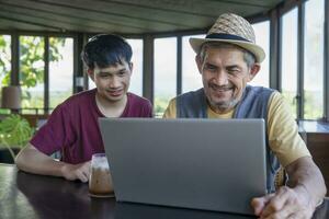 mature homme sur vacances prend une café Pause avec le sien fils dans café sur le montagne, destination dans Thaïlande, travail plus de ordinateur portable, concept de numérique nomade, voyage travailler librement à distance sur l'Internet en ligne photo