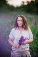 Jeune femme des stands dans blanc chemise dans champ de violet et rose lupins. magnifique Jeune femme avec frisé cheveux en plein air sur une prairie, lupins fleurir. le coucher du soleil ou lever du soleil, brillant soir lumière photo