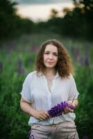 Jeune femme des stands dans blanc chemise dans champ de violet et rose lupins. magnifique Jeune femme avec frisé cheveux en plein air sur une prairie, lupins fleurir. le coucher du soleil ou lever du soleil, brillant soir lumière photo