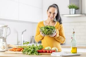 souriant femme avec une bol plein de épinard feuilles le long de avec Frais des légumes et olive pétrole dans sa cuisine photo