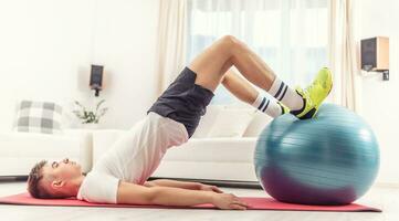 Jeune Masculin travail en dehors avec une bleu en forme Balle à Accueil sur une rouge tapis, exercice fessiers, jambes et coeur de le corps photo