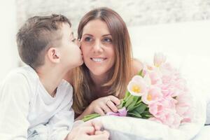 content de la mère journée concept. maman avec fils sur lit avec cadeau et tulipes.son baisers le sien maman photo