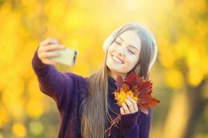 portrait de content Jeune fille avec écouteurs et téléphone intelligent dans l'automne parc écouter la musique ou fait du selfie photo