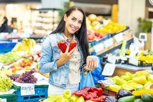 magnifique souriant femme sélectionne deux rouge poivre de une large assortiment de Frais fruit et veg dans le marché photo