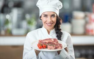 souriant femelle chef détient une assiette avec brut steaks souriant à le caméra photo