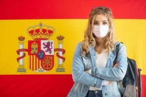 femelle étudiant transporter sac à dos des stands dans de face de le Espagnol drapeau avec une visage masque sur photo