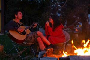 une homme pièces le guitare, une femme écoute et chante le long de. une couple dans l'amour est séance par le Extérieur feu de camp dans le Cour de le maison sur camping chaises, une romantique soir photo