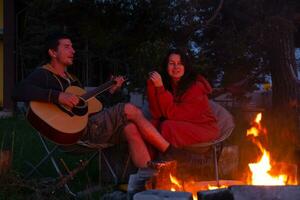 une homme pièces le guitare, une femme écoute et chante le long de. une couple dans l'amour est séance par le Extérieur feu de camp dans le Cour de le maison sur camping chaises, une romantique soir photo
