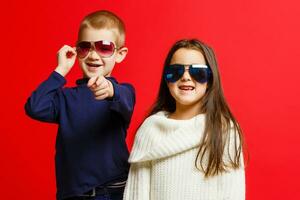 studio portrait de de bonne humeur peu fille et garçon dans des lunettes de soleil, isolé sur rouge Contexte photo