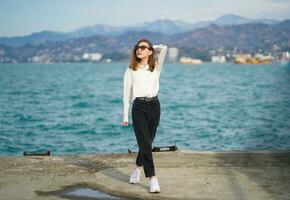 femme en marchant près mer. nature, de rechange temps concept. Jeune femme avec décontractée vêtements permanent près l'eau. paysage marin avec montagnes. marcher sur béton digue. fille portant des lunettes de soleil regards en haut dans ciel photo