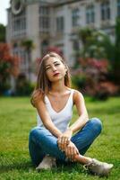 magnifique Jeune femme portant réservoir Haut et jeans séance sur herbe pelouse dans de face de bâtiment. Jeune femme posant séance sur vert herbe. ville des loisirs. insouciant la personne repos expérience calme photo