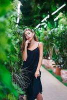 en pensant magnifique femme permanent dans fourré de tropical les plantes. Jeune femme portant noir robe sur mince les bretelles des stands parmi vert les plantes à l'intérieur serre. fille regards directement à le caméra. photo