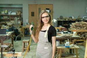 travailleur Charpentier femme en utilisant outils souriant en toute confiance Jeune femelle menuisier dans tablier permanent près Table de travail et à la recherche à caméra amical tandis que travail dans le artisanat atelier photo