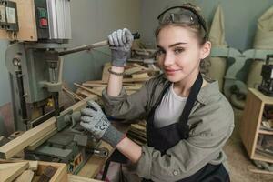 travailleur Charpentier femme en utilisant outils souriant en toute confiance Jeune femelle menuisier dans tablier permanent près Table de travail et à la recherche à caméra amical tandis que travail dans le artisanat atelier photo