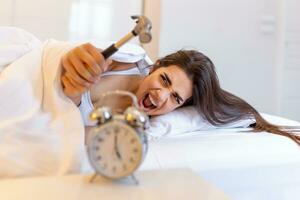 Jeune femme essaie à Pause le alarme l'horloge avec marteau, détruire le horloge. fille mensonge dans lit tournant de un alarme l'horloge avec marteau dans le Matin à 5h du matin. photo