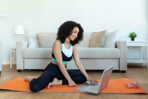 un adulte africain femme Est-ce que yoga et force formation des exercices sur une tapis dans sa vivant chambre. elle suit un en ligne exercice cours vidéo sur sa portable. photo