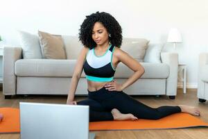 un adulte africain femme Est-ce que yoga et force formation des exercices sur une tapis dans sa vivant chambre. elle suit un en ligne exercice cours vidéo sur sa portable. photo