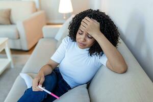 triste, inquiet africain américain femme vérification sa récent grossesse test, séance sur canapé à maison. maternité, enfant naissance et famille problèmes concept. indésirable grossesse photo