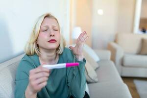 triste, inquiet femme vérification sa récent grossesse test, séance sur canapé à maison. maternité, enfant naissance et famille problèmes concept. indésirable grossesse photo