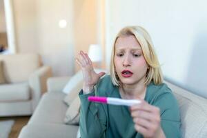 triste, inquiet femme vérification sa récent grossesse test, séance sur canapé à maison. maternité, enfant naissance et famille problèmes concept. indésirable grossesse photo