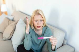 triste, inquiet femme vérification sa récent grossesse test, séance sur canapé à maison. maternité, enfant naissance et famille problèmes concept. indésirable grossesse photo