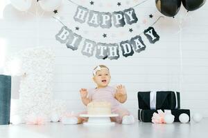 une un année vieux fille est assis sur le sol près une sucré gâteau dans honneur de sa premier anniversaire, essaie le gâteau et sourires, des rires photo