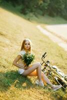 portrait de une magnifique fille dans une champ, séance sur le herbe, suivant à une vélo, avec une bouquet de verveine fleurs dans le réglage Soleil dans été photo