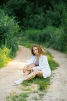 une souriant fille dans une blanc chemise est séance sur une forêt chemin dans une été vert parc photo