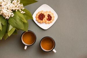 deux des tasses avec thé, biscuits sur une assiette et bouquets de fleurs de lys de le vallée sur une vert Contexte. pause pour repos, lent vie. Haut vue photo