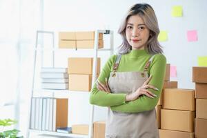 petit affaires entrepreneur PME, sourire asiatique Jeune femme, fille propriétaire permanent sur de soi avec bras franchi préparer commande envoyer à client emballage boîte à livraison, travail en ligne à maison, commerce électronique. photo