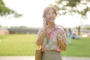 portrait de asiatique Jeune femme voyageur avec tissage panier, mobile téléphone sur vert Publique parc Contexte. périple voyage mode de vie, monde Voyage explorateur ou Asie été tourisme concept. photo