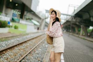 Jeune asiatique femme voyageur avec tissage panier content souriant à la recherche à une caméra à côté de train chemin de fer. périple voyage mode de vie, monde Voyage explorateur ou Asie été tourisme concept. photo