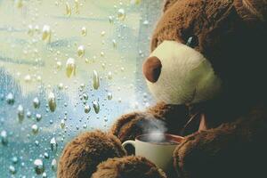 une marron jouet ours est assis sur une fenêtre avec une tasse de thé et regards à gouttes de pluie sur le verre. photo