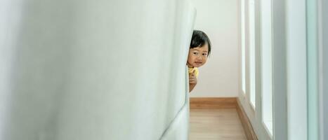 peu asiatique les enfants enfant en jouant cacher et chercher permanent derrière le canapé, adorable enfant ayant amusement dans le maison. content famille avoir amusement avec enfants, activité, apprentissage, activité, jeu, méditation photo