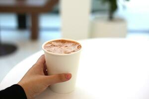cacao Lait papier tasse en portant dans femme main sur bois plateau avec blanc table dans café photo