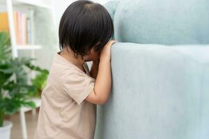 peu asiatique les enfants enfant en jouant cacher et chercher permanent derrière le canapé, adorable enfant ayant amusement dans le maison. content famille avoir amusement avec enfants, activité, apprentissage, activité, jeu, méditation photo