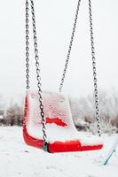 Balançoire bébé couverte de neige en hiver - aire de jeux vide - balançoire en plastique rouge dans le froid photo