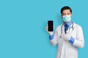 docteur tenant le téléphone montrant le pouce vers le haut isolé. médecin pakistanais sur fond bleu. écran clair du téléphone. photo