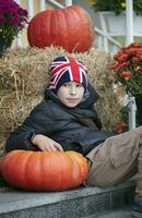 une garçon dans une chapeau avec le Anglais drapeau est assis avec grand citrouilles. photo