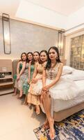 une groupe de asiatique femmes séance ensemble sur une blanc lit tandis que portant Robes et maquillage pendant une fête à une amis maison photo