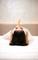 un asiatique femme dans une blanc robe est mensonge sur une blanc lit avec sa noir cheveux ample dans une Hôtel photo